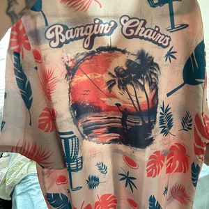 May include: A beige button-up shirt with a tropical print. The shirt features a sunset scene with palm trees, a silhouette of a person throwing a disc, and the text "Bangin' Chains".
