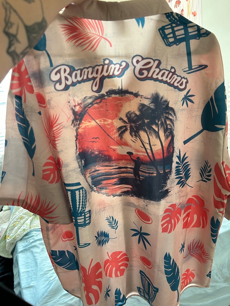 May include: A beige button-up shirt with a tropical print. The shirt features a sunset scene with palm trees, a silhouette of a person throwing a disc, and the text "Bangin' Chains".