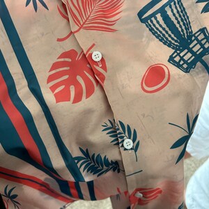 May include: A beige button-up shirt with a tropical print featuring red and blue palm leaves, a red monstera leaf, and a blue disc golf basket. The shirt has a white button closure.