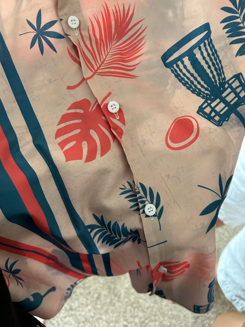 May include: A beige button-up shirt with a tropical print featuring red and blue palm leaves, a red monstera leaf, and a blue disc golf basket. The shirt has a white button closure.