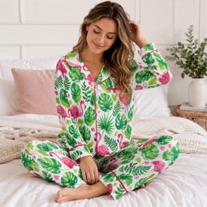 May include: A two-piece pyjama set featuring a long-sleeved button-down shirt and matching trousers. The fabric is white with a vibrant all-over print of pink flamingos and various green tropical leaves, finished with thin pink piping trim.