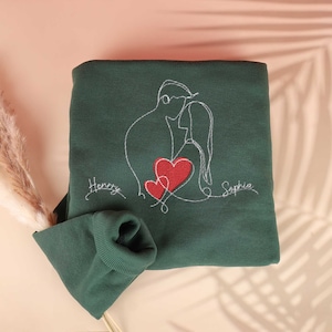 May include: A dark green sweatshirt with a white embroidered design of a couple embracing with two red hearts. The names "Henry" and "Sophia" are embroidered below the design.