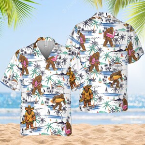 May include: White short-sleeved button-down shirt with a tropical print featuring a cartoon Bigfoot character surfing, holding a flamingo, and wearing a sombrero. The print also includes palm trees, sailboats, and beach scenes.