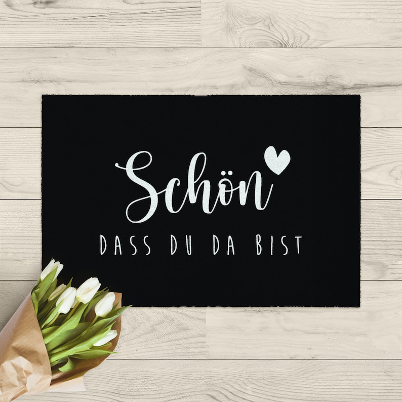 K&ouml;nnte beinhalten: Schwarze Willkommensmatte mit wei&szlig;em Text "Sch&ouml;n dass du da bist" und einem wei&szlig;en Herz.