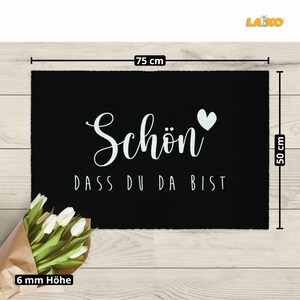 K&ouml;nnte beinhalten: Schwarze Fu&szlig;matte mit wei&szlig;em Text "Sch&ouml;n dass du da bist" und einem wei&szlig;en Herz. Die Fu&szlig;matte misst 75 cm x 50 cm und ist 6 mm dick.