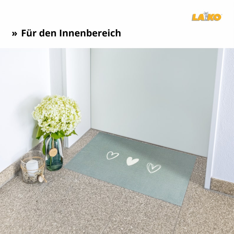 May include: A light blue doormat with three white heart designs. The doormat is placed on a tiled floor in front of a white door. A vase with white flowers is on the floor to the left of the doormat.