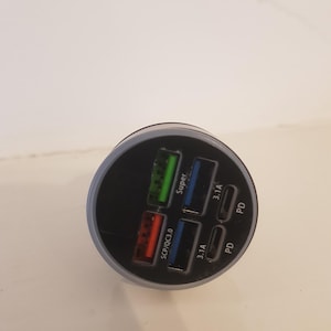 May include: A black and gray USB car charger with multiple ports. The charger has ports labeled "Super", "3.1A", "SCP/QC3.0", and "PD". The ports are colored green, blue, and red.