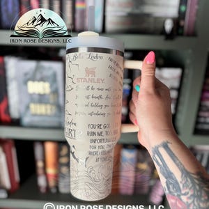 May include: A stainless steel tumbler with a white background and black and gray illustrations. The tumbler has a straw and a handle. The text on the tumbler reads "Bella Ladra", "Stanley", "You're going to ruin me, too, but unfortunately for you, that's where I feel most at home".