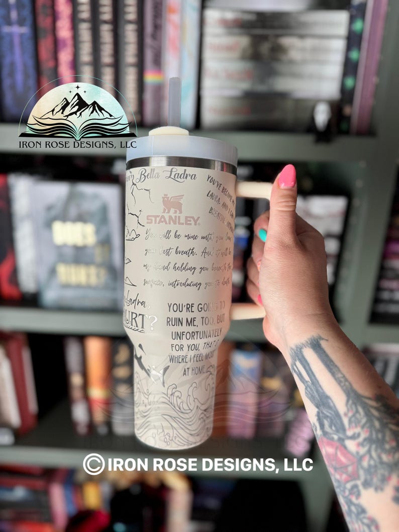 May include: A stainless steel tumbler with a white background and black and gray illustrations. The tumbler has a straw and a handle. The text on the tumbler reads "Bella Ladra", "Stanley", "You're going to ruin me, too, but unfortunately for you, that's where I feel most at home".