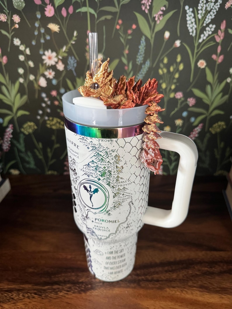 May include: White travel mug with a dragon design and a map of the fictional land of Poromiel. The mug has a black handle and a straw. The dragon is gold and red.