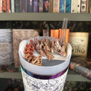 May include: A white travel mug with a map design and a gold and red dragon figurine on top. The mug has a clear lid and a straw.