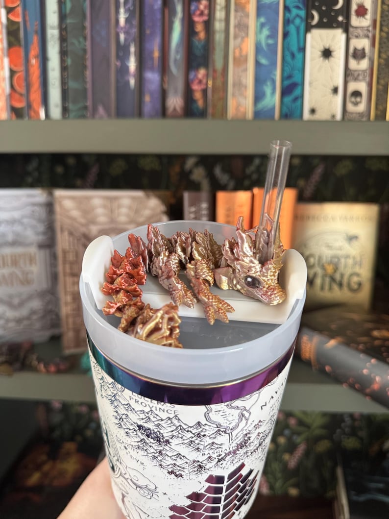 May include: A white travel mug with a map design and a gold and red dragon figurine on top. The mug has a clear lid and a straw.