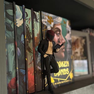 Könnte beinhalten: Eine Figur einer Person in einer schwarzen Jacke, schwarzen Hose und einem roten Helm, die sich an ein Bücherregal lehnt. Die Figur hält ein Buch und steht vor mehreren Büchern mit farbenfrohen Einbänden. Die Buchumschläge haben Blumen- und Porträtmuster.