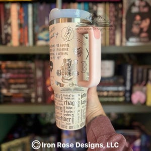May include: A beige stainless steel tumbler with a light pink handle and lid. The tumbler features various silver-colored graphics and text, including the words "Grape" and "She Kickers". The tumbler is held in front of a bookshelf.