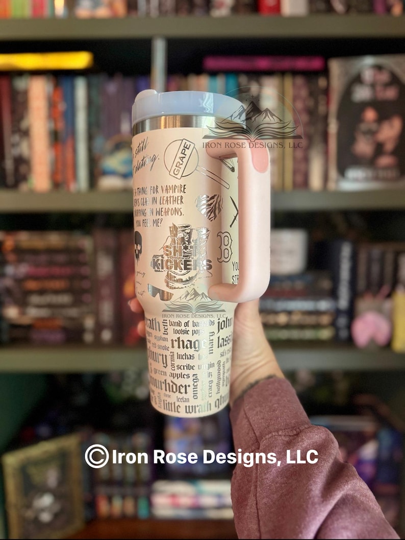 May include: A beige stainless steel tumbler with a light pink handle and lid. The tumbler features various silver-colored graphics and text, including the words "Grape" and "She Kickers". The tumbler is held in front of a bookshelf.