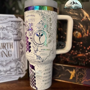 May include: White travel mug with a purple and white dragon design. The mug features a map of a fictional land with the words "Navarre", "Morraine Province", "Elsum Province", "Brae Province", "Poromiel", "Pavis", "Krovla Province", and "Cordyn". The mug also has the text "I am the storm and the fury of every story that has ever been written".