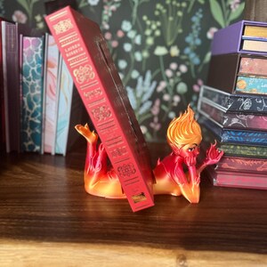 K&ouml;nnte beinhalten: Ein rotes und orangefarbenes Buchst&uuml;tze in Form einer liegenden mythologischen Figur, die ein rotes Buch h&auml;lt. Die Buchst&uuml;tze steht auf einer Holzoberfl&auml;che, mit anderen B&uuml;chern und dekorativen Gegenst&auml;nden im Hintergrund. Das Buch hat goldene Buchstaben.