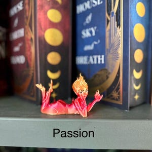 K&ouml;nnte beinhalten: Eine rot-orangefarbene Figur einer Frau mit feurigem Haar, die mit ausgestreckten Armen liegt. Das Wort "Passion" ist in Schwarz auf dem Regal darunter aufgedruckt. Im Hintergrund sind zwei dekorative B&uuml;cher zu sehen.