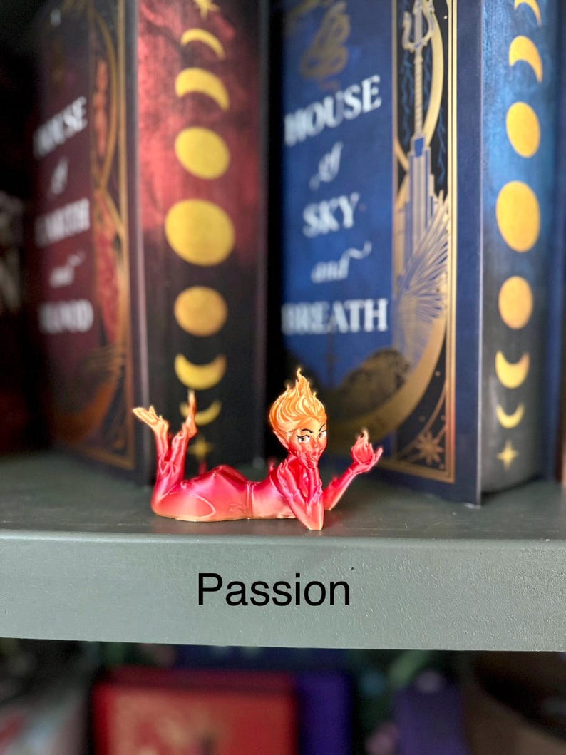 K&ouml;nnte beinhalten: Eine rot-orangefarbene Figur einer Frau mit feurigem Haar, die mit ausgestreckten Armen liegt. Das Wort "Passion" ist in Schwarz auf dem Regal darunter aufgedruckt. Im Hintergrund sind zwei dekorative B&uuml;cher zu sehen.