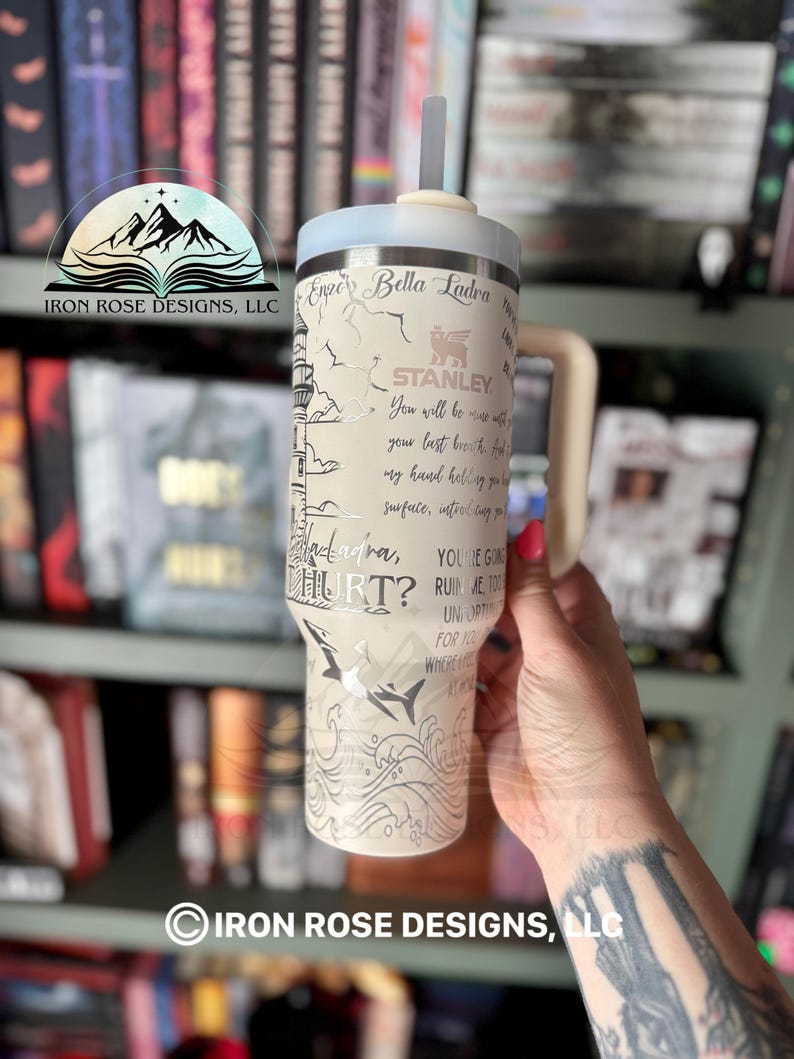 May include: A white stainless steel tumbler with a black and white graphic of a lighthouse and waves. The tumbler has a straw and a lid. The text "Enzo Bella Ladra" and "You're going to ruin me too" is printed on the tumbler.