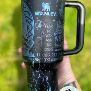May include: A black stainless steel tumbler with a blue iridescent lid and handle. The tumbler features a graphic design with a skull, a flower, and the text "I fear no gods and no kings. He is order. She is chaos. One cannot exist without the other." The tumbler is made by Stanley.