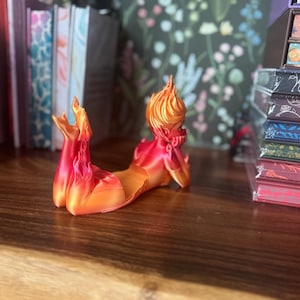 K&ouml;nnte beinhalten: Eine 3D-gedruckte Figur einer liegenden Figur mit einem flammenartigen Kopf und K&ouml;rper. Die Figur ist haupts&auml;chlich rot und orange, mit einer glatten, gl&auml;nzenden Oberfl&auml;che. Sie liegt auf einer Holzoberfl&auml;che, mit B&uuml;chern und dekorativen Gegenst&auml;nden im Hintergrund.
