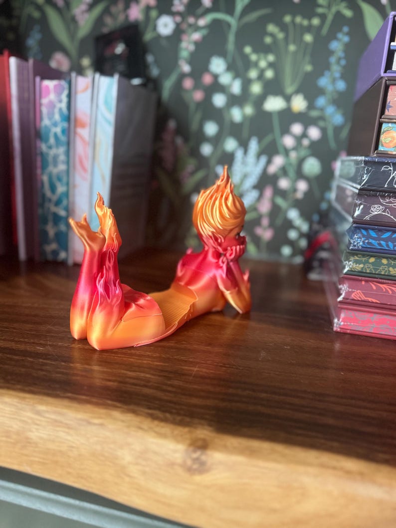 K&ouml;nnte beinhalten: Eine 3D-gedruckte Figur einer liegenden Figur mit einem flammenartigen Kopf und K&ouml;rper. Die Figur ist haupts&auml;chlich rot und orange, mit einer glatten, gl&auml;nzenden Oberfl&auml;che. Sie liegt auf einer Holzoberfl&auml;che, mit B&uuml;chern und dekorativen Gegenst&auml;nden im Hintergrund.