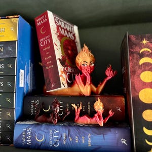 K&ouml;nnte beinhalten: Ein B&uuml;cherregal mit drei Fantasy-Romanen und dekorativen Buchst&uuml;tzen. Die B&uuml;cher tragen die Titel "Crescent City" und "House of Earth and Blood" von Sarah J. Maas. Die Buchst&uuml;tzen haben die Form einer feurigen weiblichen Figur.