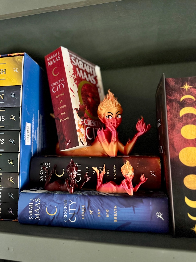 K&ouml;nnte beinhalten: Ein B&uuml;cherregal mit drei Fantasy-Romanen und dekorativen Buchst&uuml;tzen. Die B&uuml;cher tragen die Titel "Crescent City" und "House of Earth and Blood" von Sarah J. Maas. Die Buchst&uuml;tzen haben die Form einer feurigen weiblichen Figur.