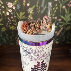 May include: A white stainless steel tumbler with a map of a fictional land printed on it. The tumbler has a iridescent purple and blue band around the middle. A small dragon figurine with gold and brown accents is attached to the lid, and a clear drinking straw is sticking out of the lid.