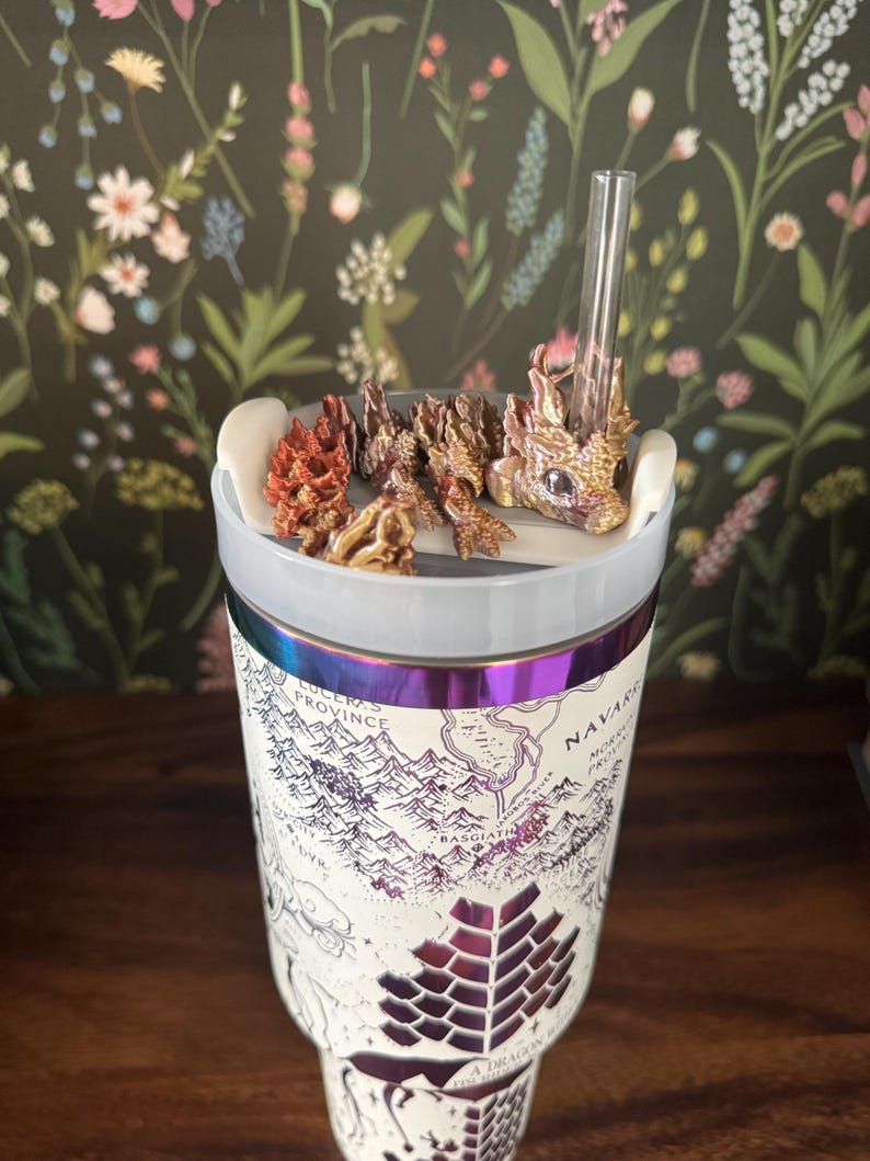 May include: A white stainless steel tumbler with a map of a fictional land printed on it. The tumbler has a iridescent purple and blue band around the middle. A small dragon figurine with gold and brown accents is attached to the lid, and a clear drinking straw is sticking out of the lid.