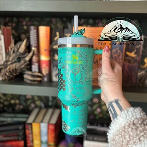 May include: A turquoise stainless steel tumbler with a gold design featuring a dragon, honeycomb pattern, and the text "FOROMIEL" and "WINGED ACE". The tumbler has a lid and a straw.