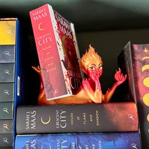 May include: A vibrant, orange and red bookend shaped like a mythical creature, holding a book titled "Crescent City" by Sarah J. Maas. The bookend sits on a shelf with other books from the same series, featuring dark covers with gold lettering.