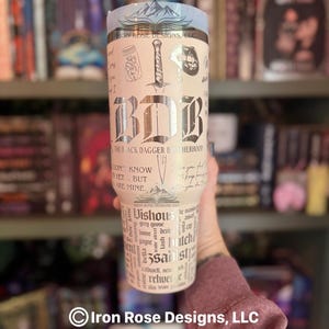 May include: A stainless steel tumbler with a light blue top and a cream-colored body. The tumbler features the text "BDB" in a gothic font, along with illustrations and text from "The Black Dagger Brotherhood" series. The tumbler is held in a person's hand.