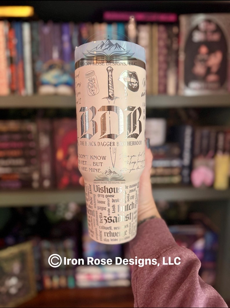 May include: A stainless steel tumbler with a light blue top and a cream-colored body. The tumbler features the text "BDB" in a gothic font, along with illustrations and text from "The Black Dagger Brotherhood" series. The tumbler is held in a person's hand.