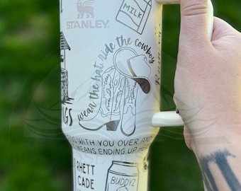 May include: White stainless steel tumbler with a black and white design featuring a cowboy boot, a hat, a milk carton, a building, and the text "wear the hat ride the cowboy". The tumbler has a white lid and a white handle. The text "BUDDYZ BEST" is also visible on the tumbler.