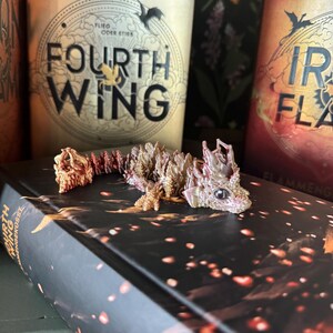 May include: A small, pink and orange dragon figurine with a textured, scaly body. The dragon is lying on a black book with the title "Fourth Wing" and the author "Rebecca Yarros" printed on the cover. The book is on a green surface.