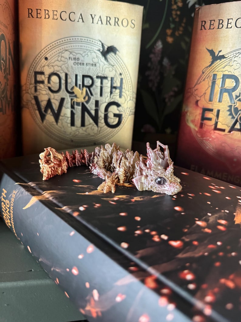 May include: A small, pink and orange dragon figurine with a textured, scaly body. The dragon is lying on a black book with the title "Fourth Wing" and the author "Rebecca Yarros" printed on the cover. The book is on a green surface.