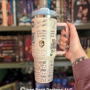 May include: A cream-colored tumbler with a light blue lid and handle, featuring various phrases and graphics in black. The tumbler includes the text "We still ain't dating" and "I have a thing for vampire".