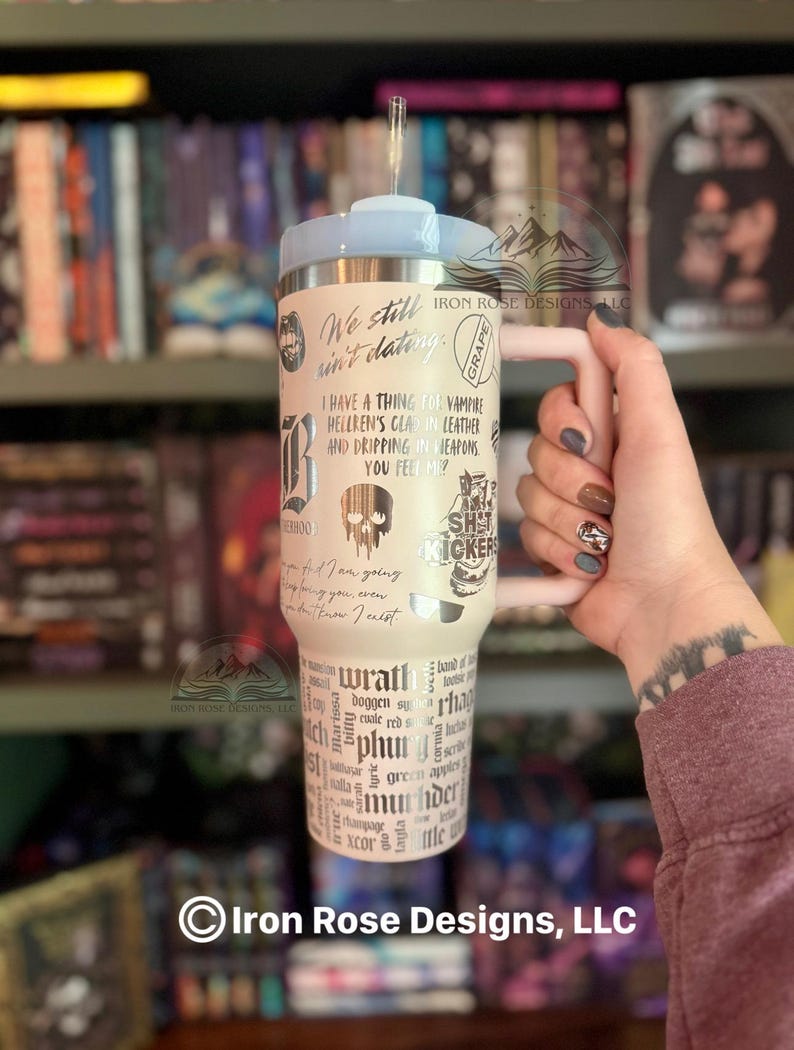 May include: A cream-colored tumbler with a light blue lid and handle, featuring various phrases and graphics in black. The tumbler includes the text "We still ain't dating" and "I have a thing for vampire".
