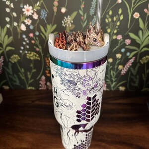 May include: A white stainless steel tumbler with a map design and a dragon figurine on top. The tumbler has a clear lid and a straw. The map design includes the words "Lucus Province" and "Bassiant".