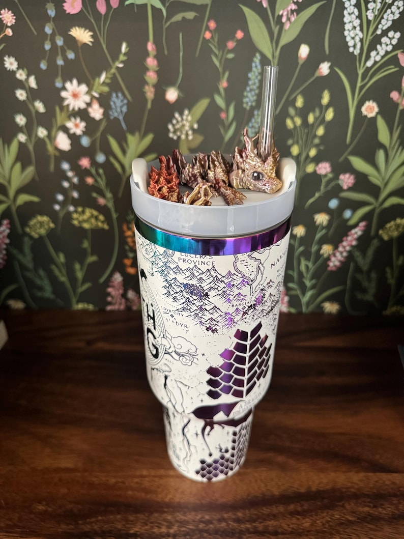 May include: A white stainless steel tumbler with a map design and a dragon figurine on top. The tumbler has a clear lid and a straw. The map design includes the words "Lucus Province" and "Bassiant".