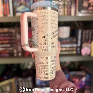 May include: A beige travel mug with a light pink handle and lid. The mug features etched text and graphics, including crossed swords and phrases like "You are both my strength and my weakness."