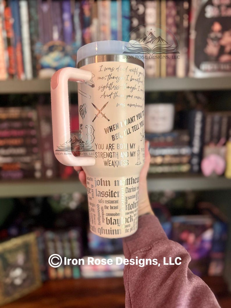 May include: A beige travel mug with a light pink handle and lid. The mug features etched text and graphics, including crossed swords and phrases like "You are both my strength and my weakness."