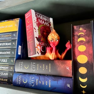 K&ouml;nnte beinhalten: Ein B&uuml;cherregal mit mehreren B&uuml;chern von Sarah J. Maas, darunter die "Crescent City"-Titel. Zwischen den B&uuml;chern befindet sich eine dekorative Figur einer feurigen Frau. Die B&uuml;cher sind in verschiedenen Farben, darunter Rot, Blau und Gelb.