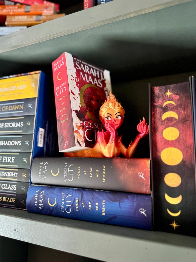 K&ouml;nnte beinhalten: Ein B&uuml;cherregal mit mehreren B&uuml;chern von Sarah J. Maas, darunter die "Crescent City"-Titel. Zwischen den B&uuml;chern befindet sich eine dekorative Figur einer feurigen Frau. Die B&uuml;cher sind in verschiedenen Farben, darunter Rot, Blau und Gelb.