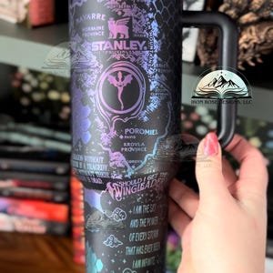 May include: A black stainless steel travel mug with a colorful iridescent finish. The mug features a map of the fictional world of the Wheel of Time book series, with text that reads "Should I get the Wingleader?" and "I am the sky and the power of every storm that has ever been. I am infinite."