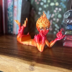 K&ouml;nnte beinhalten: 3D-gedruckte Figur einer Feuer-Elementar-Frau, in feurigen Orange- und Rott&ouml;nen. Die Figur liegt ausgestreckt, mit Flammen f&uuml;r Haare und H&auml;nde. Die Figur ruht auf einer Holzoberfl&auml;che.
