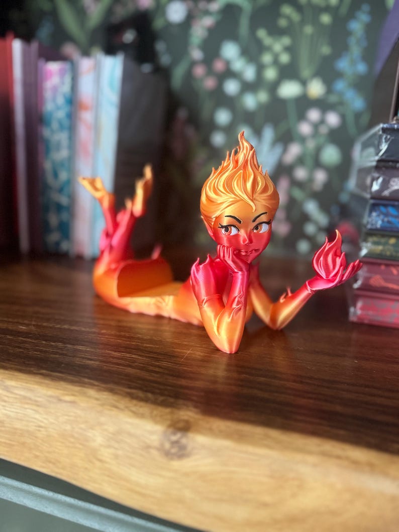 K&ouml;nnte beinhalten: 3D-gedruckte Figur einer Feuer-Elementar-Frau, in feurigen Orange- und Rott&ouml;nen. Die Figur liegt ausgestreckt, mit Flammen f&uuml;r Haare und H&auml;nde. Die Figur ruht auf einer Holzoberfl&auml;che.