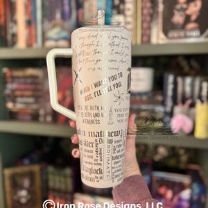 May include: A white tumbler with a large handle, featuring black text and graphics. The tumbler has quotes and words like "When I want you to beg, I'll tell you." and "John Matthew". The background is a bookshelf.